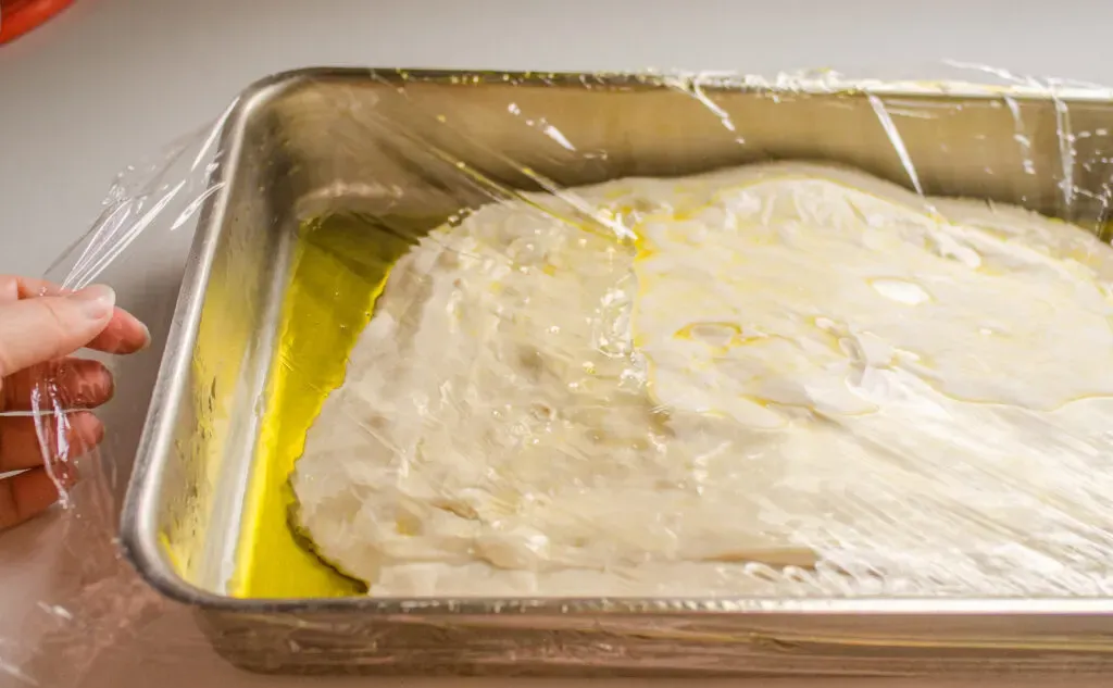 The sourdough focaccia rising in the pan with plastic wrap on top.' title='Sourdough Focaccia Recipe