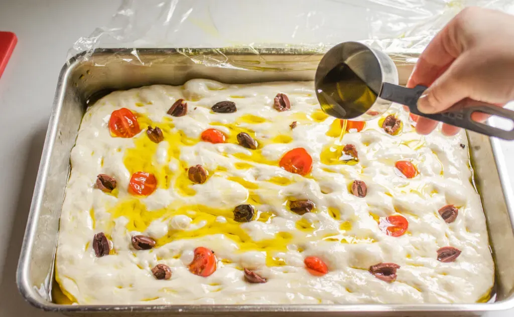 Pouring extra virgin olive oil over the toppings on the focaccia dough.' title='Sourdough Focaccia Recipe