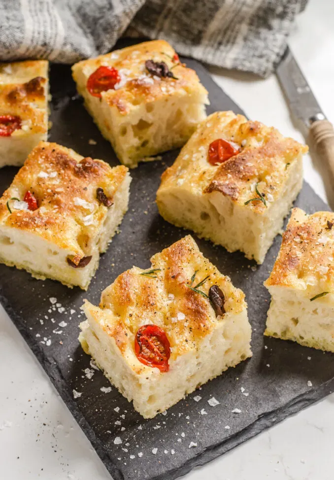 Pieces of sourdough focaccia on a slate board with a linen tea towel in the background and a serrated knife off to the side.' title='Sourdough Focaccia Recipe