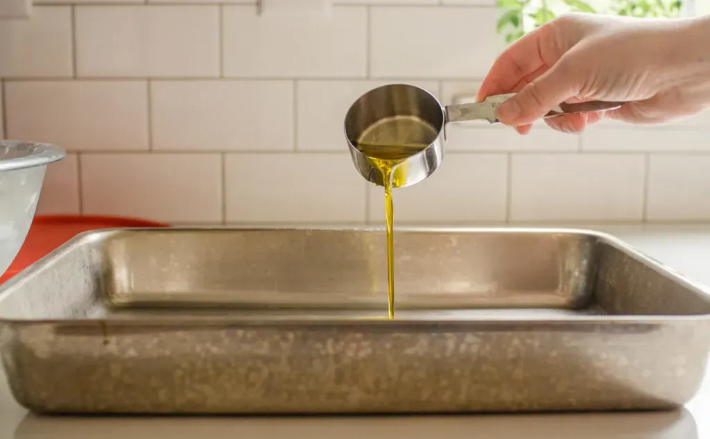 Pouring extra virgin olive oil into a pan to grease it in preparation for putting the focaccia dough in it.' title='Sourdough Focaccia Recipe