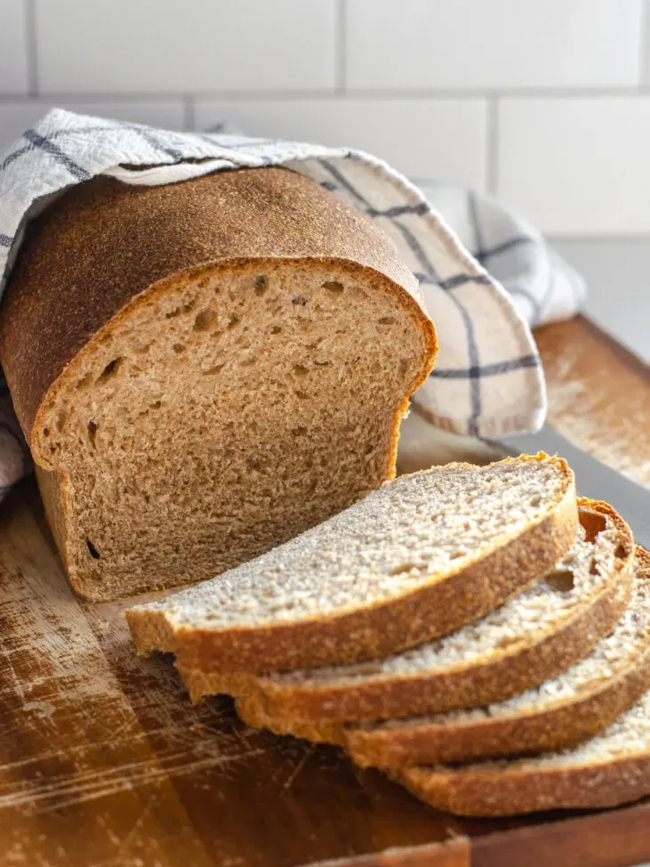 A loaf of healthy sourdough bread on a cutting board with a few slices in the foreground with the loaf in the background with a tea towel on top.' title='Healthy Whole Wheat Sourdough Sandwich Bread