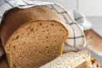 A loaf of healthy sourdough bread on a cutting board with a few slices in the foreground with the loaf in the background with a tea towel on top.' title='Healthy Whole Wheat Sourdough Sandwich Bread