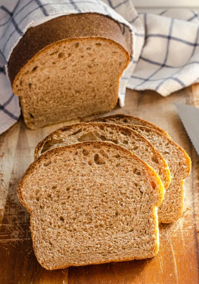 A loaf of healthy whole wheat sourdough sandwich bread on a cutting board with a few slices in the foreground with the loaf in the background with a tea towel on top.' title='Healthy Whole Wheat Sourdough Sandwich Bread