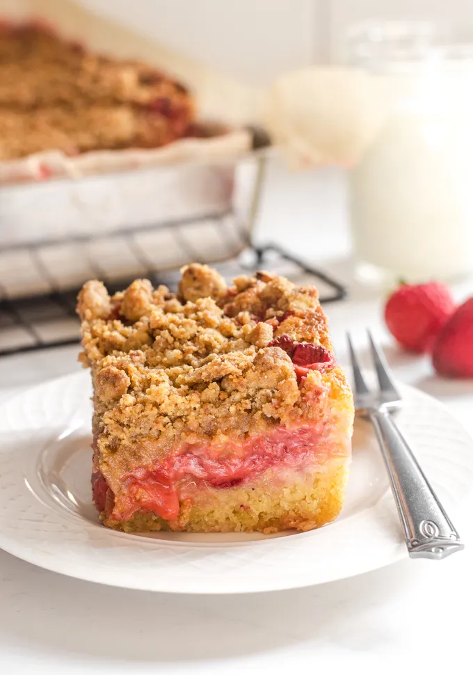 A slice of strawberry rhubarb crumb cake with strawberries and a glass of milk in the background.' title='Strawberry Rhubarb Crumb Cake