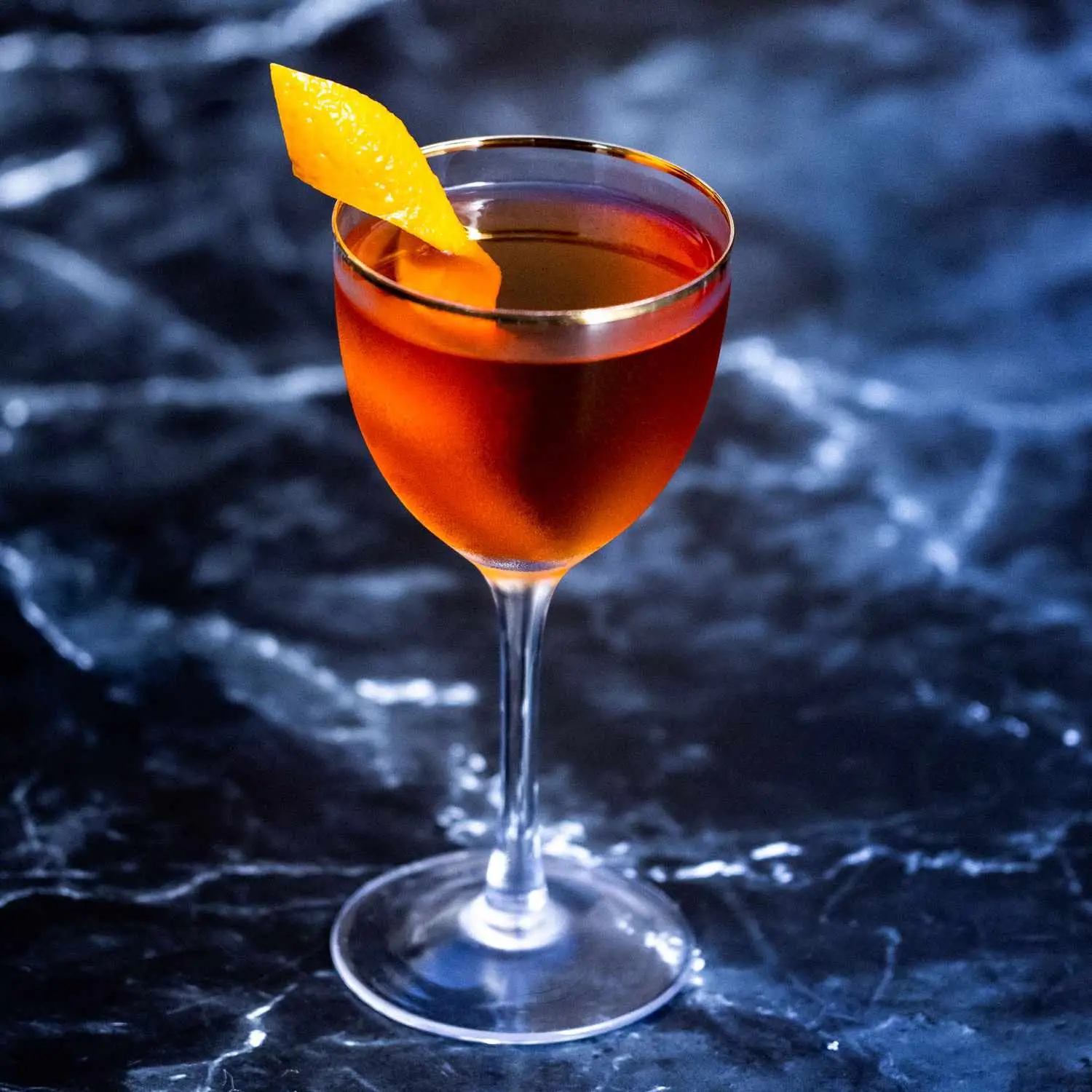 Bay Ridge cocktail in a gold-rimmed Nick & Nora glass, garnished with an orange twist, on a dark marble background