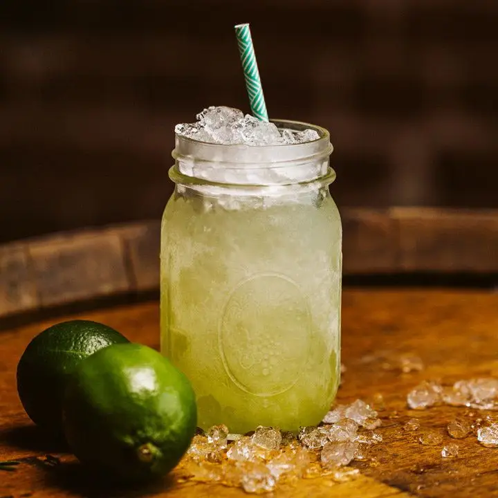 A Mason jar with a light green cocktail, crushed ice and a paper straw with limes and more crushed ice placed beside it