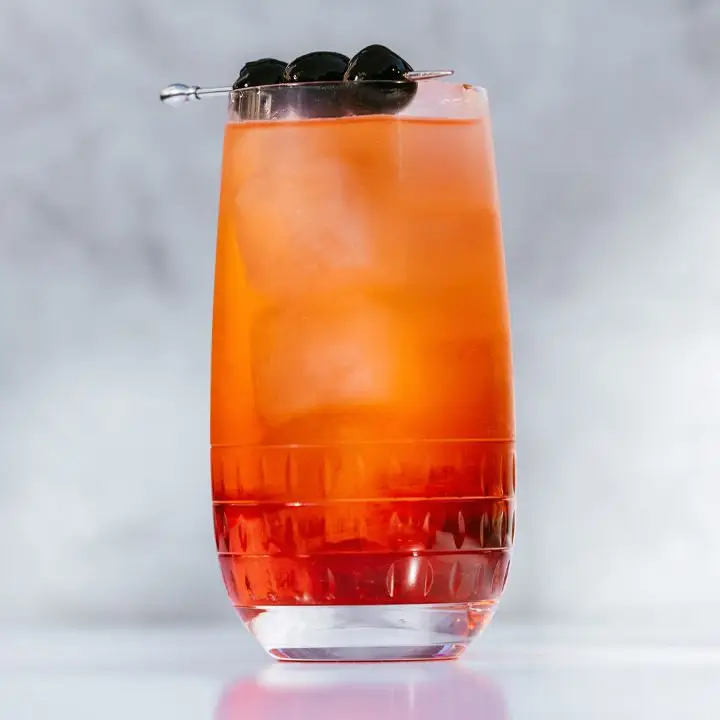 Shirley Temple in a rounded highball glass. The drink has only been lightly stirred so the red color is more intense at the base of the drink. A small metal skewer holds three Luxardo cherries and sits across the lip of the glass
