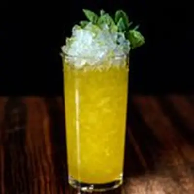 A tall Collins glass is filled with crushed ice and a golden drink, and is garnished with mint leaves. It rests on a dark wooden surface.