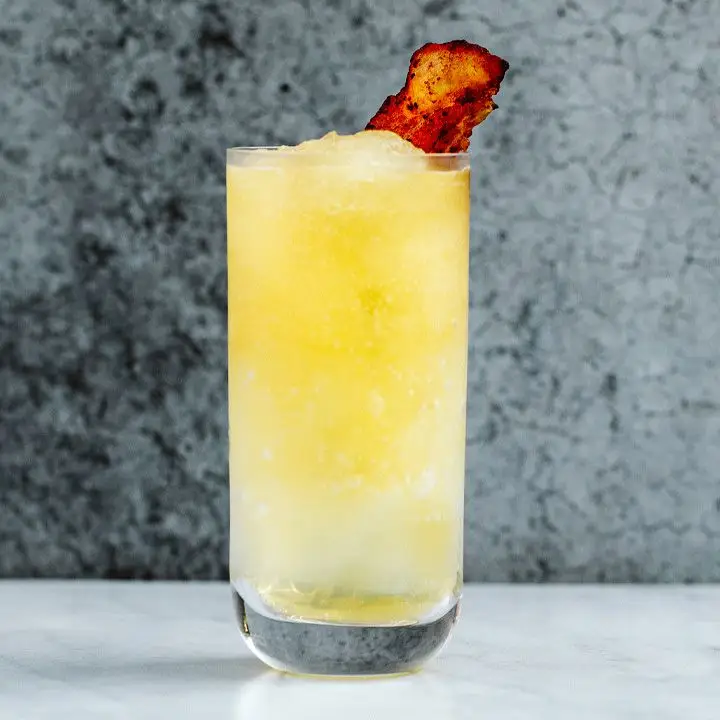 A clear Collins glass filled with a bright yellow slushie and garnished with crisp bacon