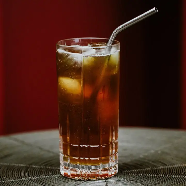 A Collins glass with a metal straw rests on a ringed mat. The drink within has a few ice cubes and is dark brown.