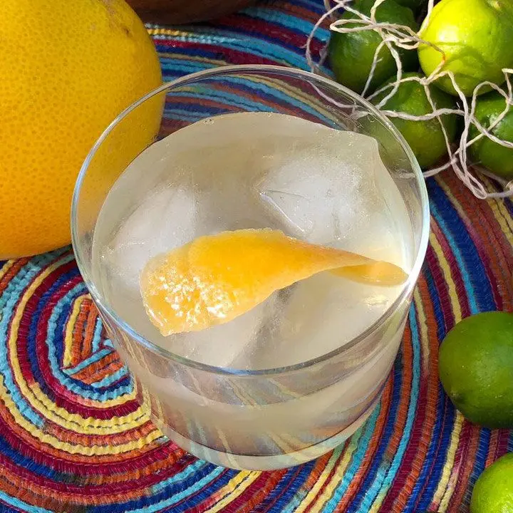 clairin regal sour cocktail in a rocks glass over ice with a grapefruit peel, served on a multi-colored mat beside key limes