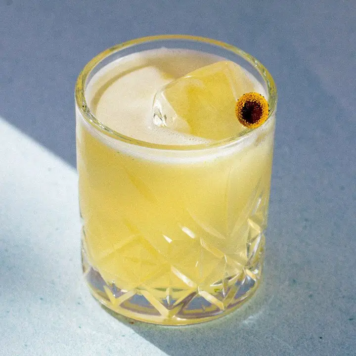 A soft yellow cocktail in a rocks glass garnished with a Szechuan button