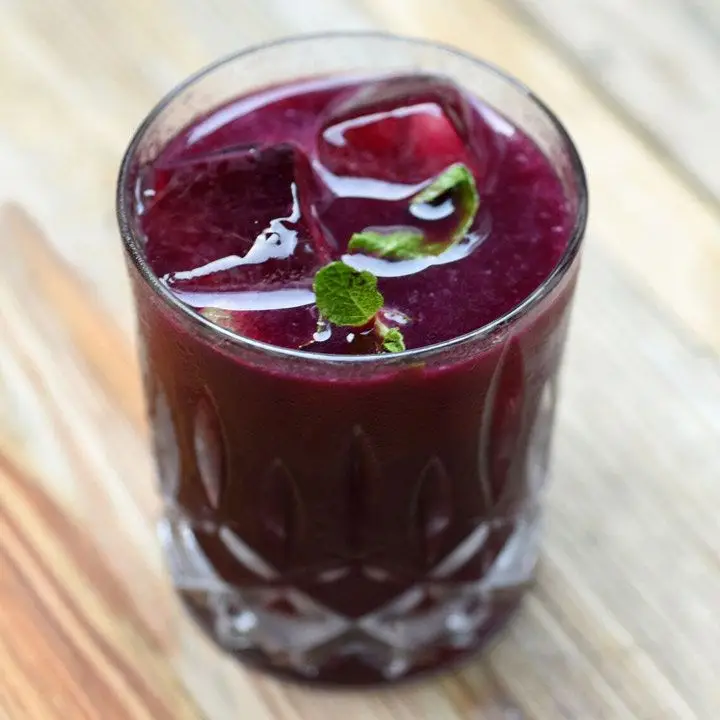 A faceted rocks glass holds a bright purple drink garnished with a few small mint leaves. The glass is resting on a wooden surface.