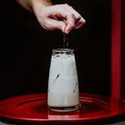 A creamy white cocktail in a tapered, beaker-like glass rests on a wide red plate. A hand garnishes it with cinnamon-sugar.