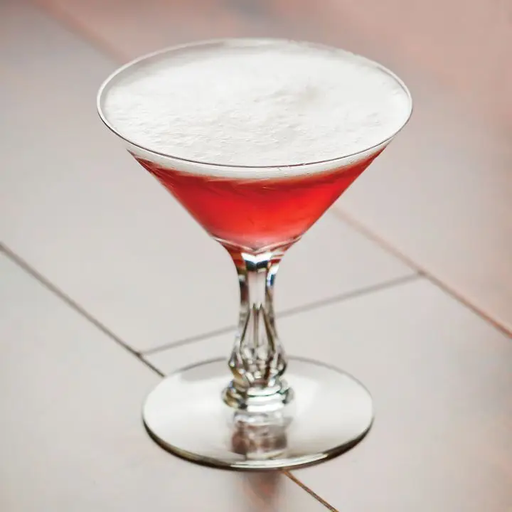 A Martini glass with a fancy stem sits on white tile. The drink is vivid red with a layer of white foam on top.