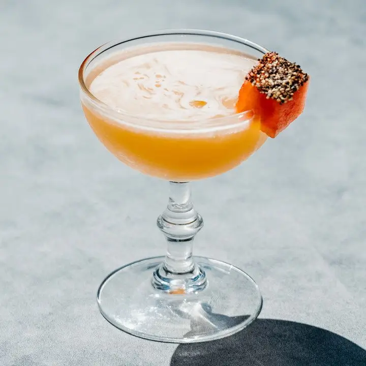 A coupe glass with a knobbed stem rests on a gray concrete surface. The drink is bright orange and garnished with a salt-and-pepper-coated chunk of watermelon.
