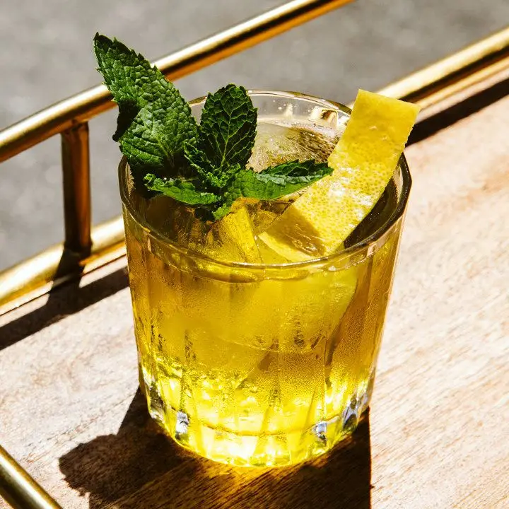 An elegant, faceted rocks glass holds a vividly golden elixir with large cubes of ice. A single trimmed lemon peel and a small mint sprig garnish the drink, which rests on a wooden bar tray.