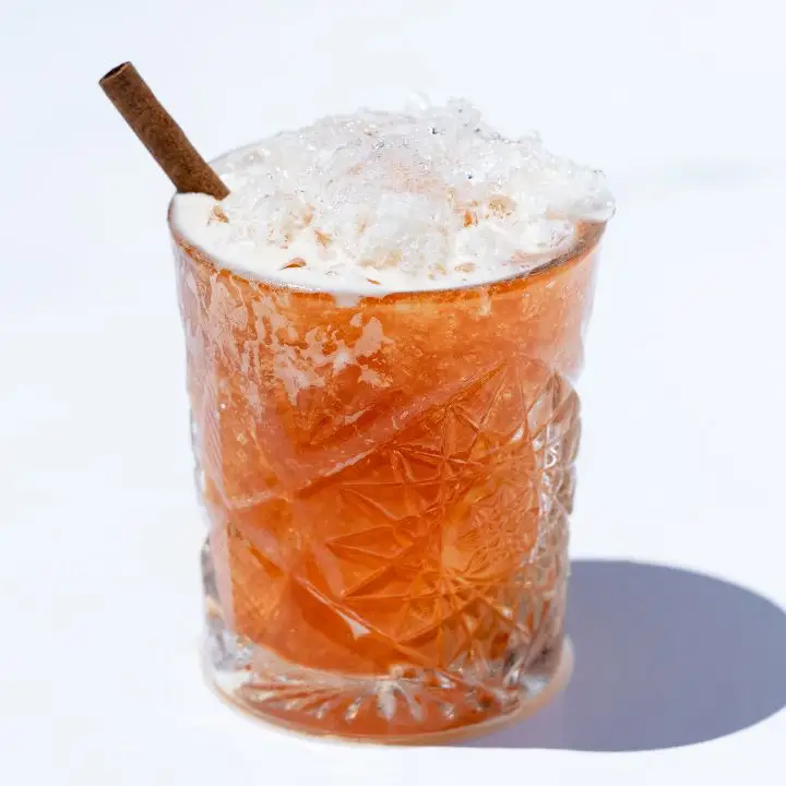 A detailed rocks glass on a stark white background holds a golden brown drink over crushed ice. It’s garnished with a cinnamon stick.