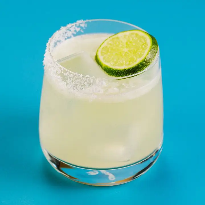 A rocks glass that tapers towards the top holds a Margarita. Half its rim is coated with salt and the other side has a lime wheel garnish. The background is solid blue. 