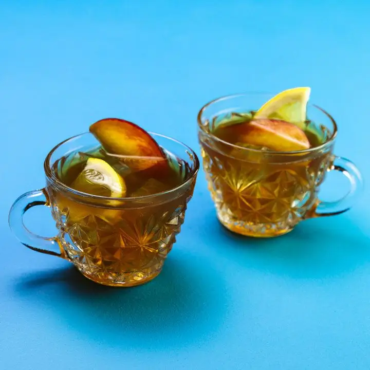 tea for two cocktail served in two ornate punch glasses, each garnished with a lemon half-wheel and peach slice