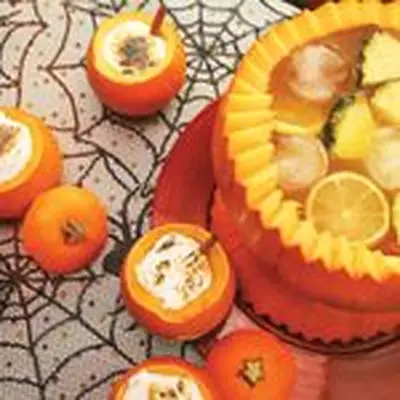 A halved pumpkin on a spider-web table cloth is filled with pineapples, oranges, ice and punch. It’s surrounded by smaller pumpkins garnished with toasted mallow and cinnamon sticks.