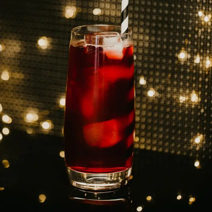 In a tapered Collins glass a few ice cubes float amongst a deep red tea. A black and white striped paper straw emerges from the the glass, and the background has a panel and glowing lights. 