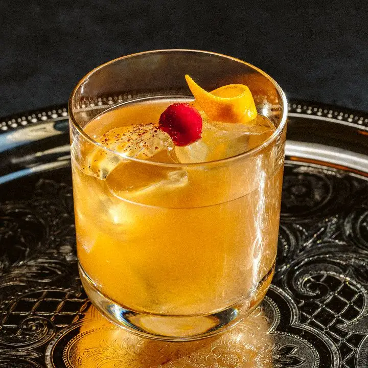 A rocks glass rests on a silver platter. The glass holds a golden drink over ice cubes, garnished with cinnamon, a single cranberry and a thin orange peel.