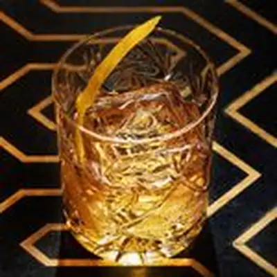 An intricately designed double Old Fashioned glass rests on a black surface with golden zig zagging inlays. The glass holds a large ice cube, a light brown drink, and a lemon zest.