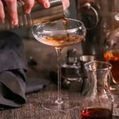 A hand pouring a Manhattan into a long-stemmed coupe glass from a mixing tin.