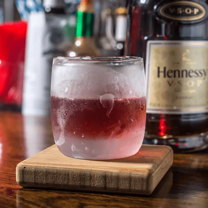A frosty rocks glass filled with a crimson hued Sazerac rests on a thick wooden coaster. A bottle of Hennessy VSOP is in the background.