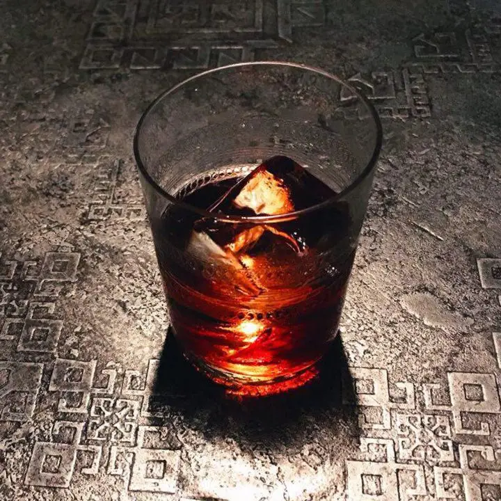 A tall rocks glass with a red drink over a single cube rests on a shiny stone surface with geometric symbols. 