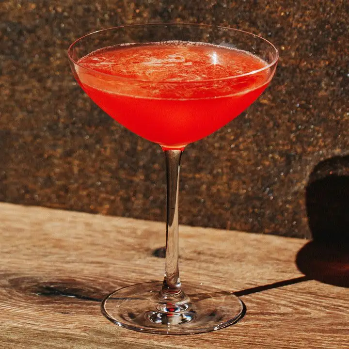 A bright red shaken cocktail in a coupe glass against a brown sparkly background