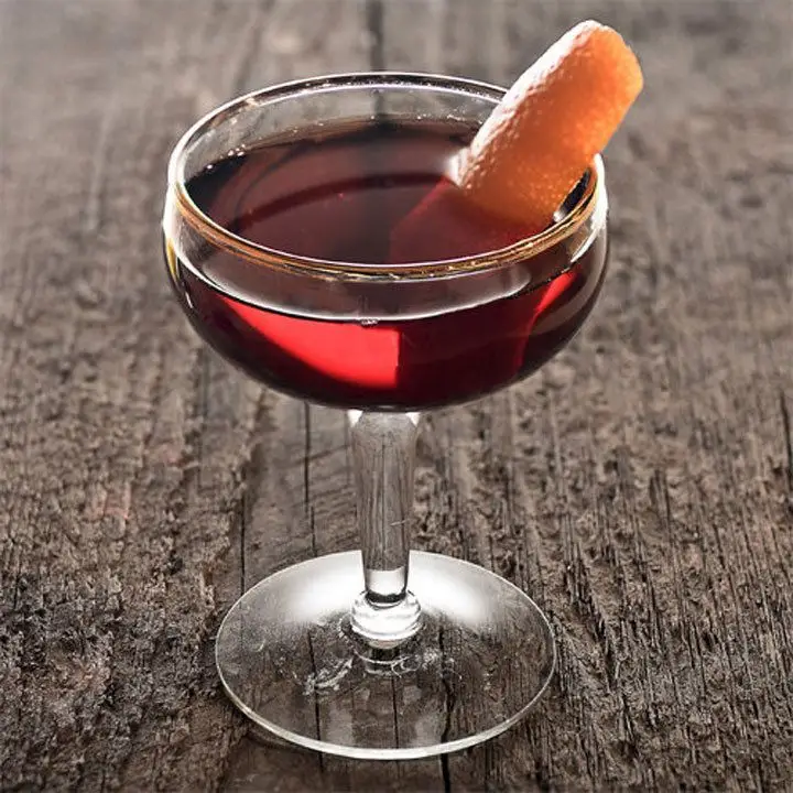 A short, unadorned coupe stands on a wooden surface. The glass holds a crimson cocktail and a thin slice of orange.
