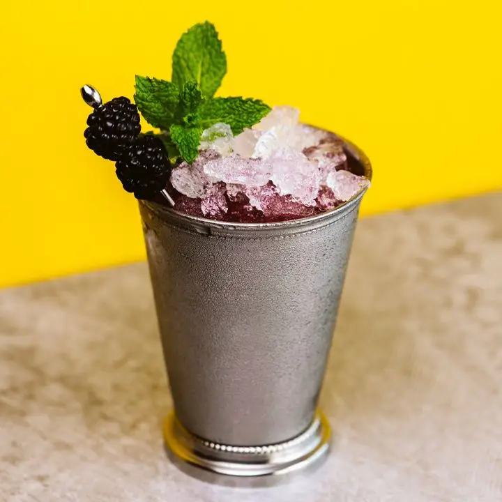A silver julep cup is beaded with condensation. The cup holds a violet liquid, a pile of crushed ice, a sprig of mint, and two blackberries pierced on a silver pick. The drink sits on a tan surface with a bright yellow wall behind it. 
