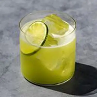 A thin-walled rocks glass sits on a gray concrete surface. The glass holds a vivid green cocktail over ice, garnished with a lime wheel.