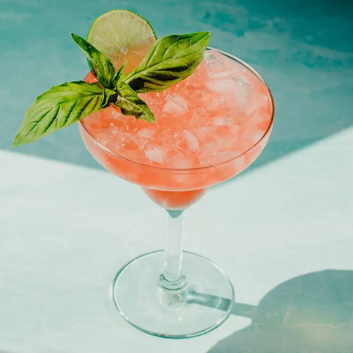 On a dramatically lit blue marble surface rests a margarita glass. It’s filled with crushed ice and a orange-flushed margarita, and garnished with a large sprig of basil and a lime wheel.