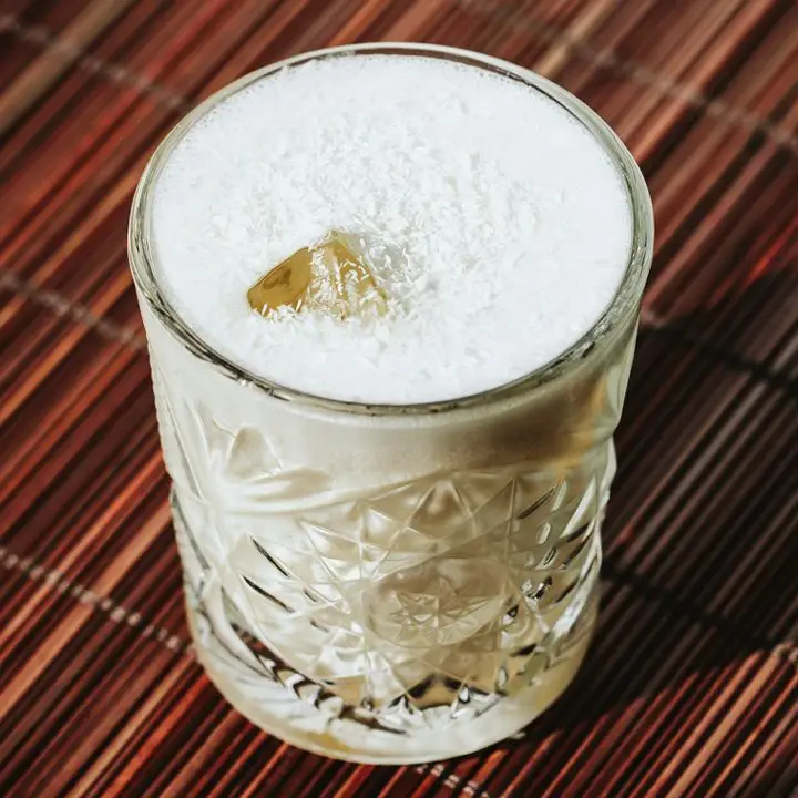 A detailed rocks glass rests on a bamboo placemat. The drink within is white, frothy and garnished with coconut flakes.