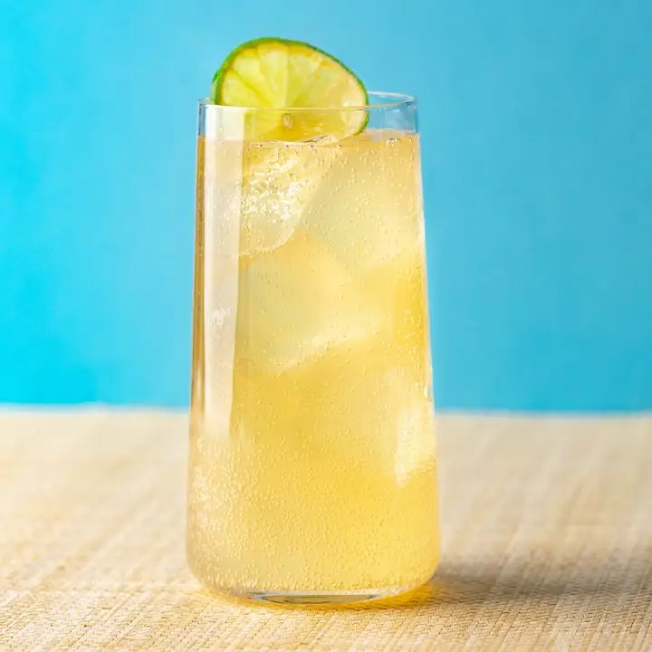 A tapered Collins glass sits on a pale blonde placement. The glass is filled with a light golden sparkling beverage over ice and garnished with a lime wheel. The background is sky blue.