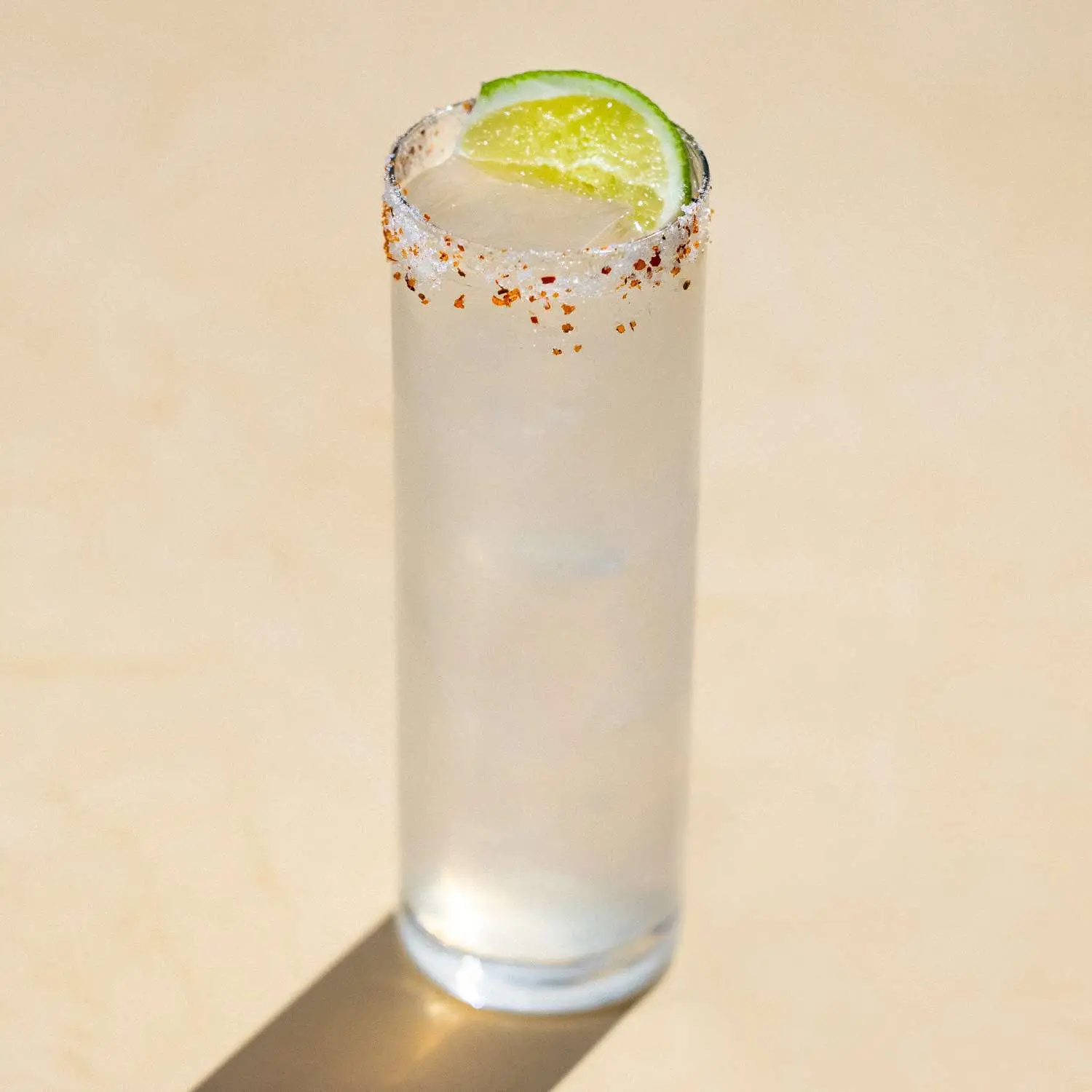 Mezcal Paloma in highball glass with Tajin salt rim and lime wedge, shot against tan background