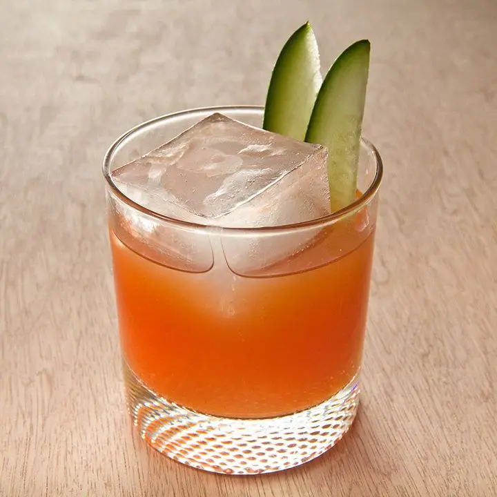 A rocks glass sits on a light colored wooden surface. In it is a bright orange tomato drink, a single large ice cube, and to slivers of cucumber.
