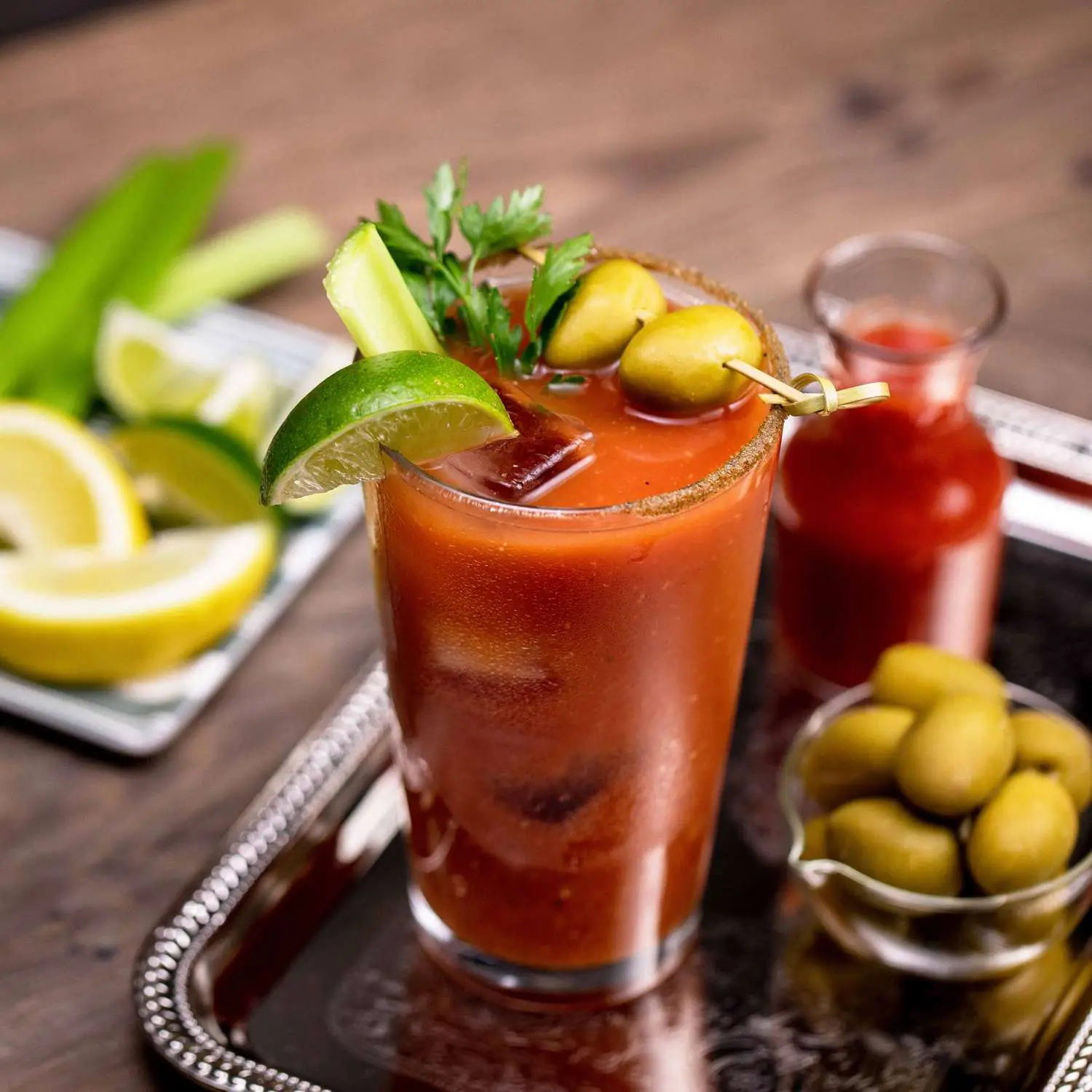 Bloody Mary in pint glass garnished with limes, parsley and olives, on silver tray surrounded by other garnishes
