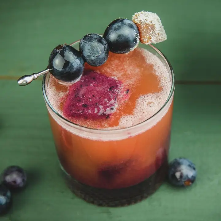 Transfusion cocktail in a rocks glass with ice cubes made from grape juice and garnished with three grapes and candied ginger on a cocktail pick.