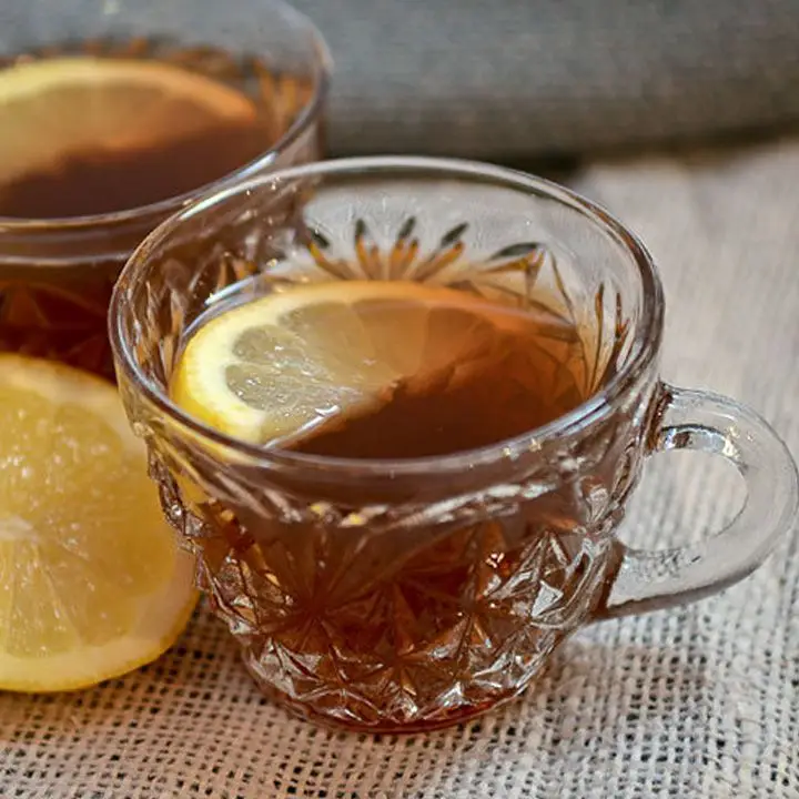 Two little glass mugs with small handles sit on burlap next to a lemon wedge. Both glasses are filled with a dark punch and garnished with a lemon slice. 