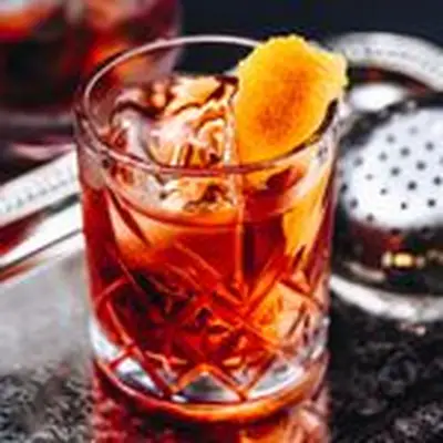 A faceted rocks glass holds a crimson-colored Negroni, a few large ice cubes, and a swath of orange peel. It sits enticingly on a black, detailed bar top