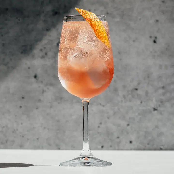 A tall wine glass stands on a white surface against a concrete backdrop. The glass is filled with a rose-colored sparkling drink, ice cubes and an orange peel.