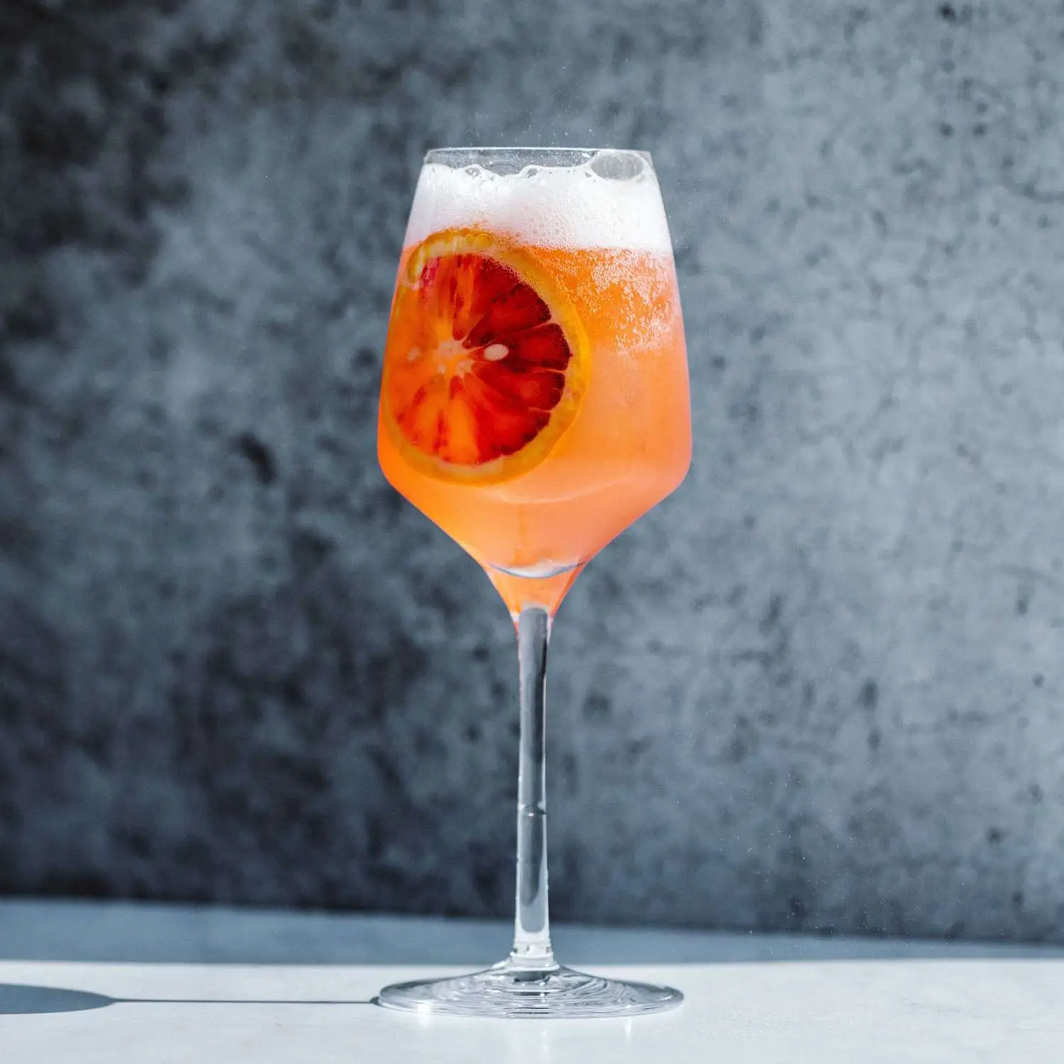 Colletti Royale pink bubbly spritz cocktail served in wine glass with blood orange wheel, shot on marble surface against gray background 