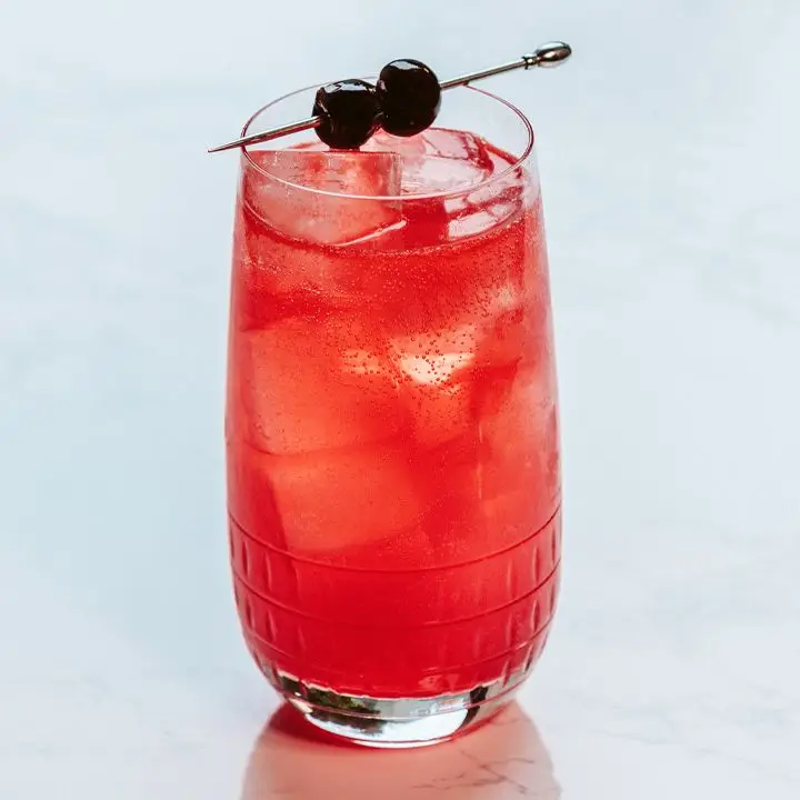 red-colored Don’t Call me Shirley in a Collins glass with two skewered cherries balanced on the rim, served on a white surface