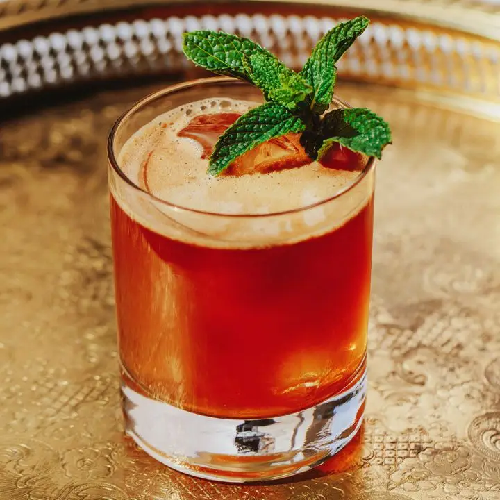 An unadorned rocks glass rests on an embossed silver platter. The drink within is dark gold and lightly foamy, with a mint sprig for garnish.