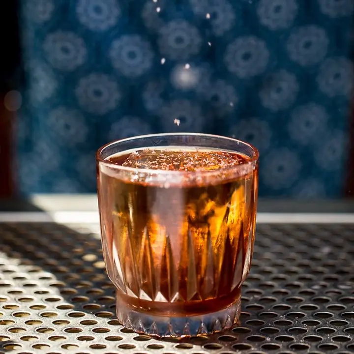 An elegant, faceted Old Fashioned glass sits on a perforated sheet of metal behind a bar. The drink within is red-gold, and a large ice cube is garnished with granulated grasshopper salt. A few flakes of the salt fall from above.