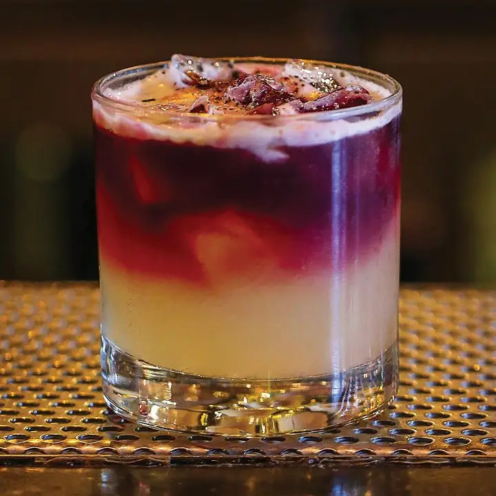 A short rocks glass rests on a bar top upon a perforated metal grate. The drink within is light gold and topped with a crimson wine that slowly gently with the drink. It’s garnished with black pepper. 
